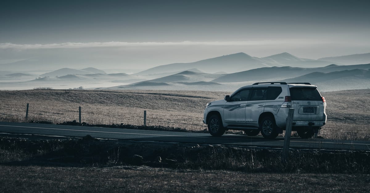 A white SUV parked on a scenic road with a backdrop of rolling hills and mountains under a cloudy sky.