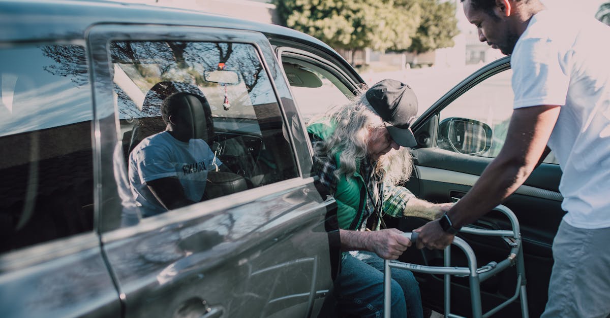 Two men help an elderly person with a walker get out of a car outdoors.